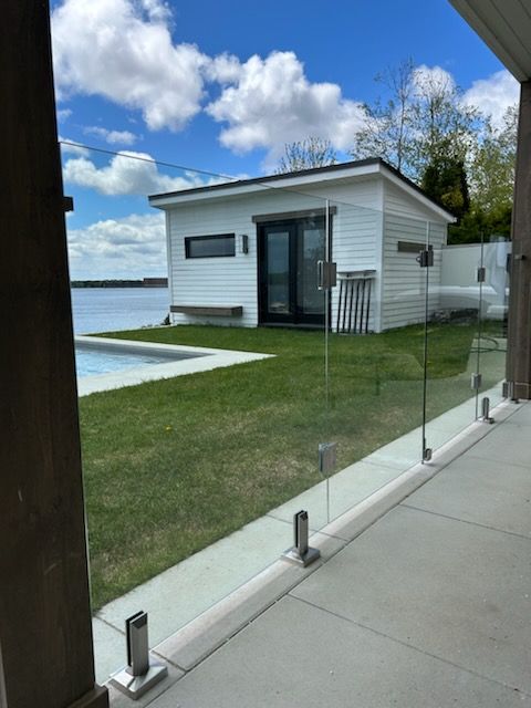 Abri blanc au bord d'une piscine, balustrade en verre sur un patio, pelouse, eau et ciel bleu.