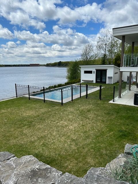 Jardin avec piscine et vue sur le lac. Herbe verte, clôture noire autour de la piscine. Bâtiment blanc. Ciel bleu, nuages.