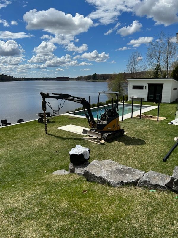 Excavatrice jaune forant près d'une piscine avec vue sur le lac, ciel bleu et bâtiment blanc.