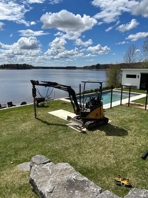Mini-pelle forant sur une pelouse verte, lac et piscine en arrière-plan sous un ciel bleu.