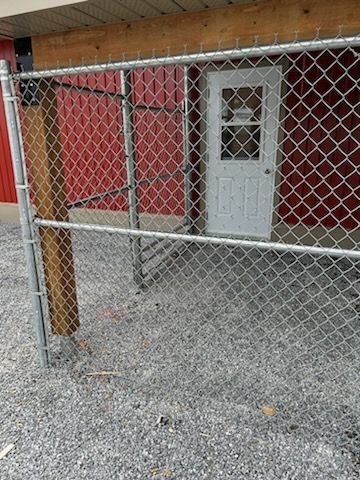 Clôture grillagée devant un bâtiment rouge avec une porte blanche. Le sol est recouvert de gravier gris.
