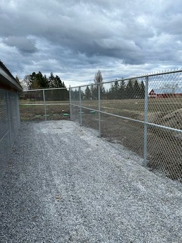 Une allée de gravier longeant une clôture grillagée. Ciel couvert, arbres au loin.