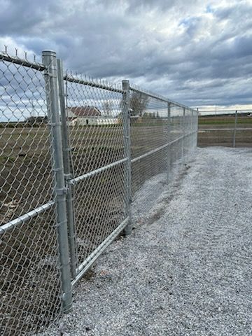 Clôture grillagée avec portail, posée sur du gravier, sous un ciel couvert.
