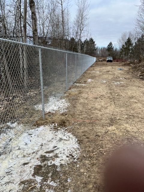Clôture grillagée longeant un chemin de terre, s'étendant jusqu'à une zone boisée. Un véhicule est visible au loin.