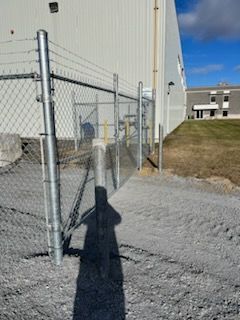 Clôture grillagée avec fil barbelé près d'un bâtiment. Ombre visible. Sol en gravier.