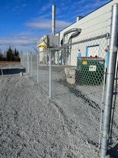 Une clôture grillagée entoure un bâtiment avec une benne verte. Sol en gravier gris. Journée ensoleillée.