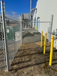 Clôture grillagée avec un portail, des bornes jaunes et un bâtiment en arrière-plan.