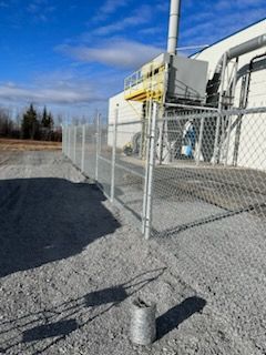 Une clôture grillagée avec un portail, près d'un bâtiment avec une haute cheminée, sur un sol de gravier sous un ciel bleu.