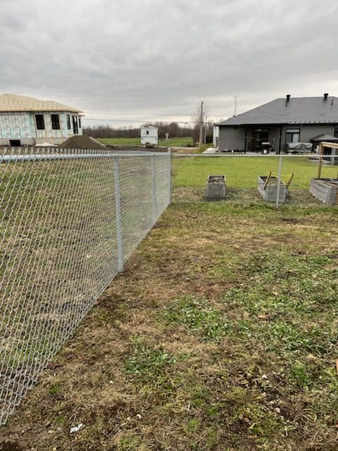 Clôture grillagée dans une cour, avec une maison en construction et une autre en arrière-plan. Ciel nuageux.