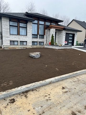Maison avec jardin fraîchement labouré, gros rocher et bordure en béton. Extérieur en pierre grise, porte de garage noire.