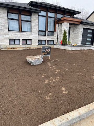 Pelouse fraîchement ensemencée devant une maison moderne ; un gros rocher repose sur le sol avec des empreintes de pas.