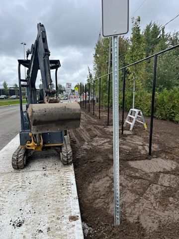 Une excavatrice creuse près d'un trottoir, près d'une clôture et d'un panneau. Ciel nuageux.