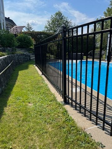 Clôture en métal noir autour d'une piscine bleue, à côté d'une zone herbeuse par une journée ensoleillée.