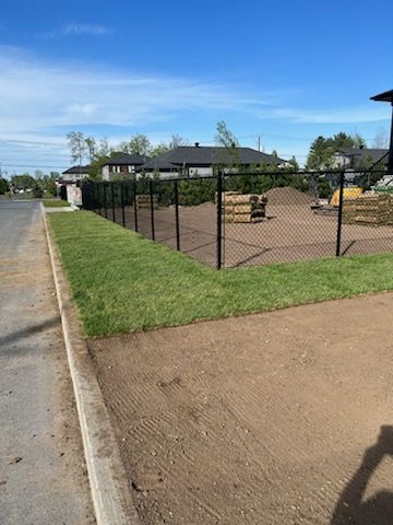 Une clôture grillagée noire entoure une zone herbeuse à côté d'un trottoir et de terre.