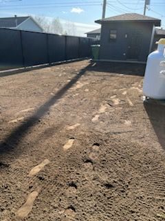 Des traces de pas sur un chemin de terre mènent à un hangar et à une clôture noire. Journée ensoleillée.