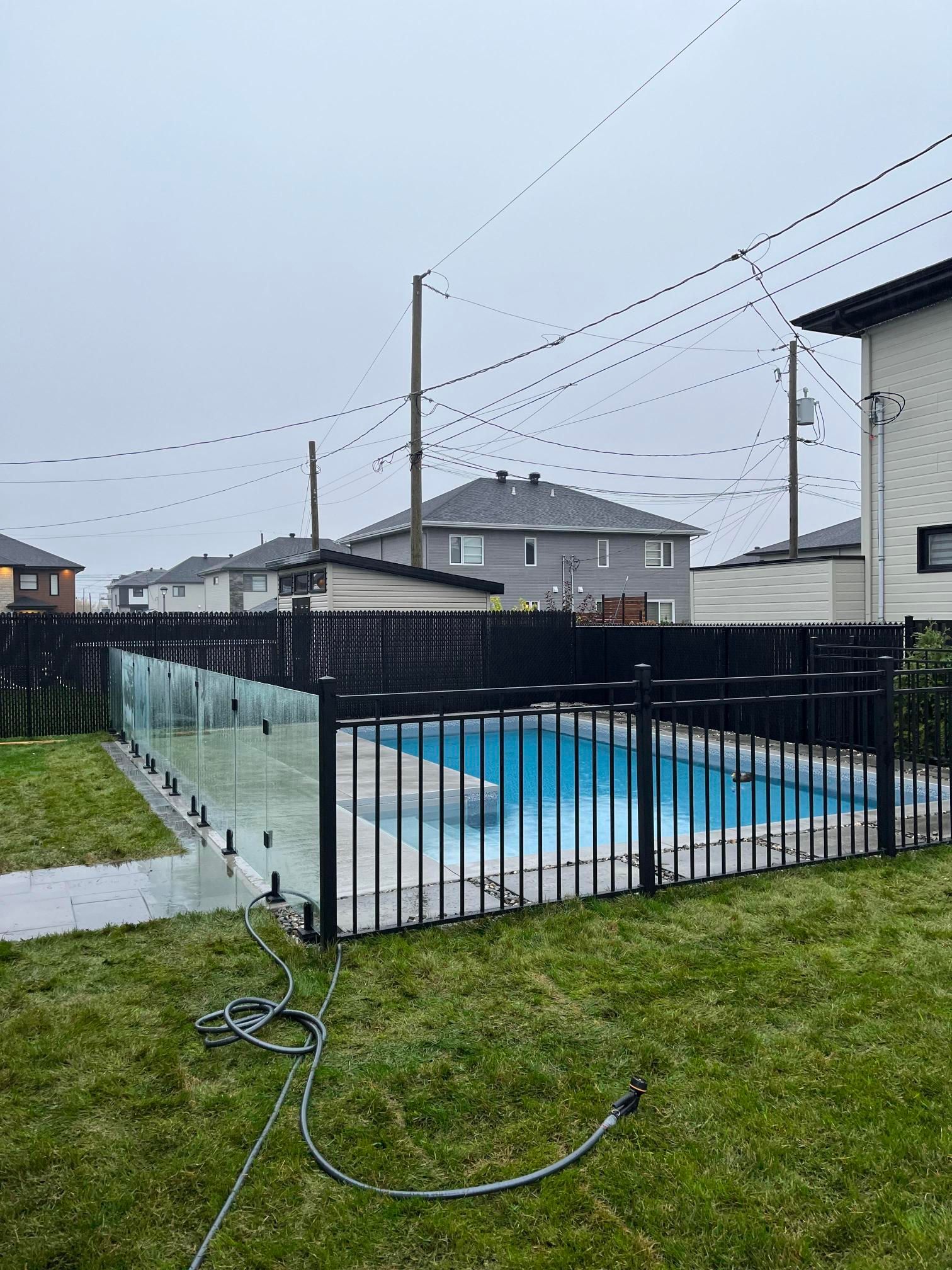 Piscine dans le jardin entourée d'une clôture en métal noir et en verre, herbe verte, ciel couvert, lignes électriques au-dessus.
