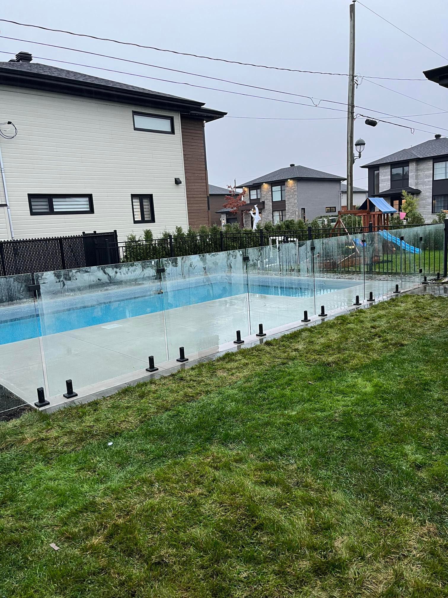 Une cour arrière avec une piscine et une clôture en verre à côté d'une maison et d'une pelouse verte.