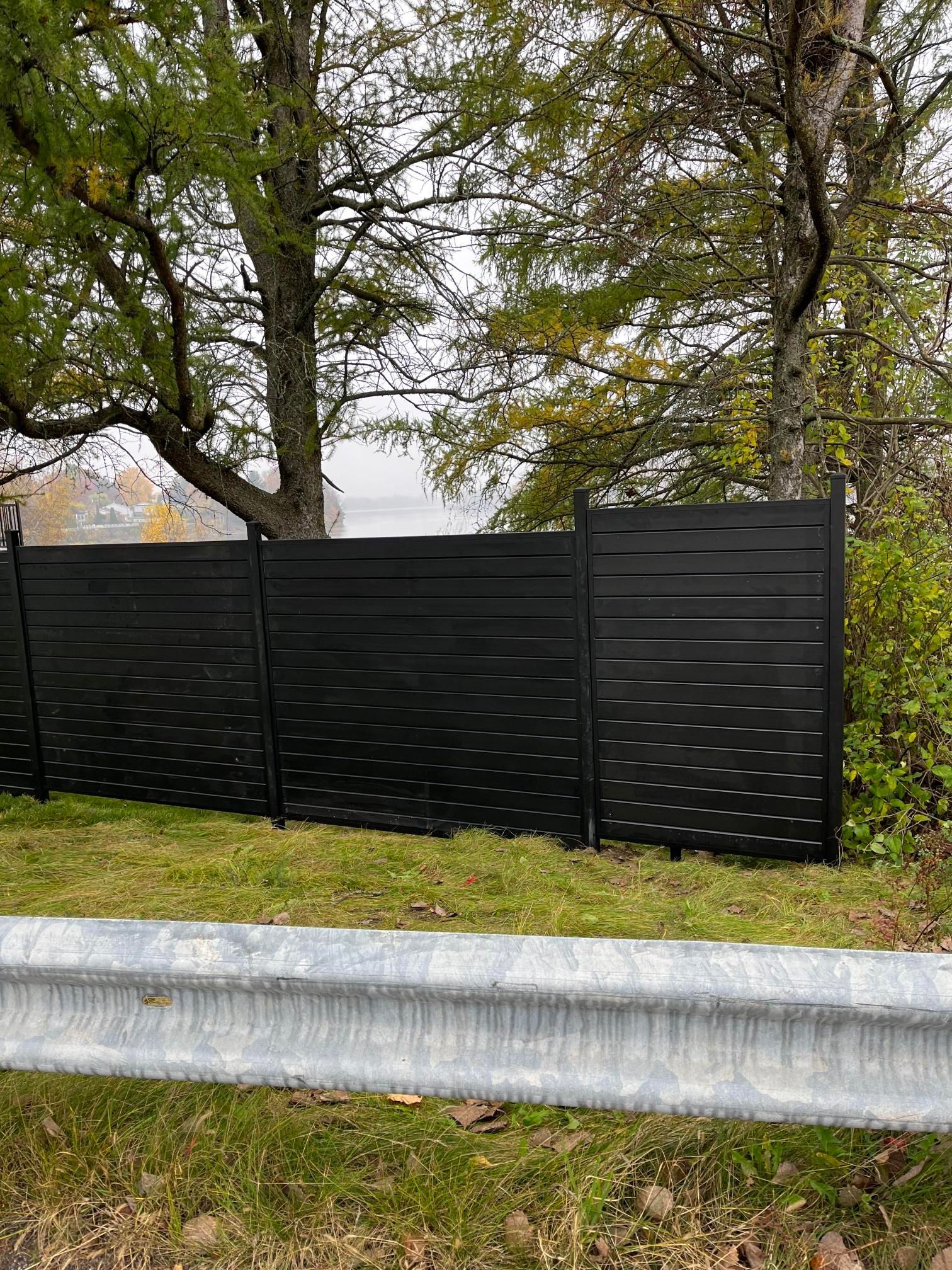 Clôture à lattes horizontales noires le long d'une route avec des arbres en arrière-plan, une barrière de sécurité en métal au premier plan.