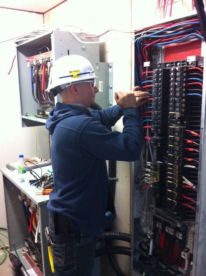 Électricien en veste bleue travaillant sur un panneau électrique, portant un casque et des lunettes de sécurité.