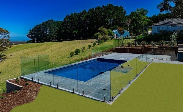 Rectangular pool with glass fence on a grassy hill, overlooking a landscape with trees and a house.