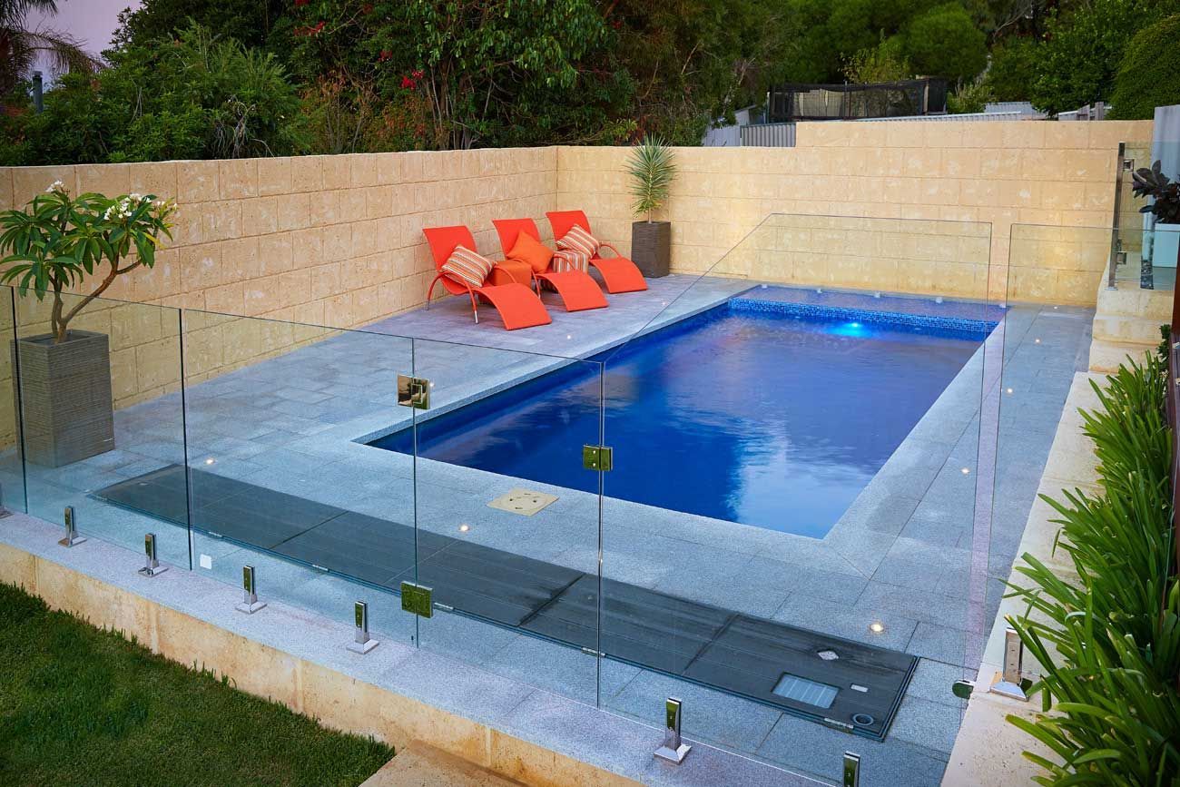 A rectangular pool with blue water, surrounded by gray paving and glass fencing, orange lounge chairs, and tan walls.
