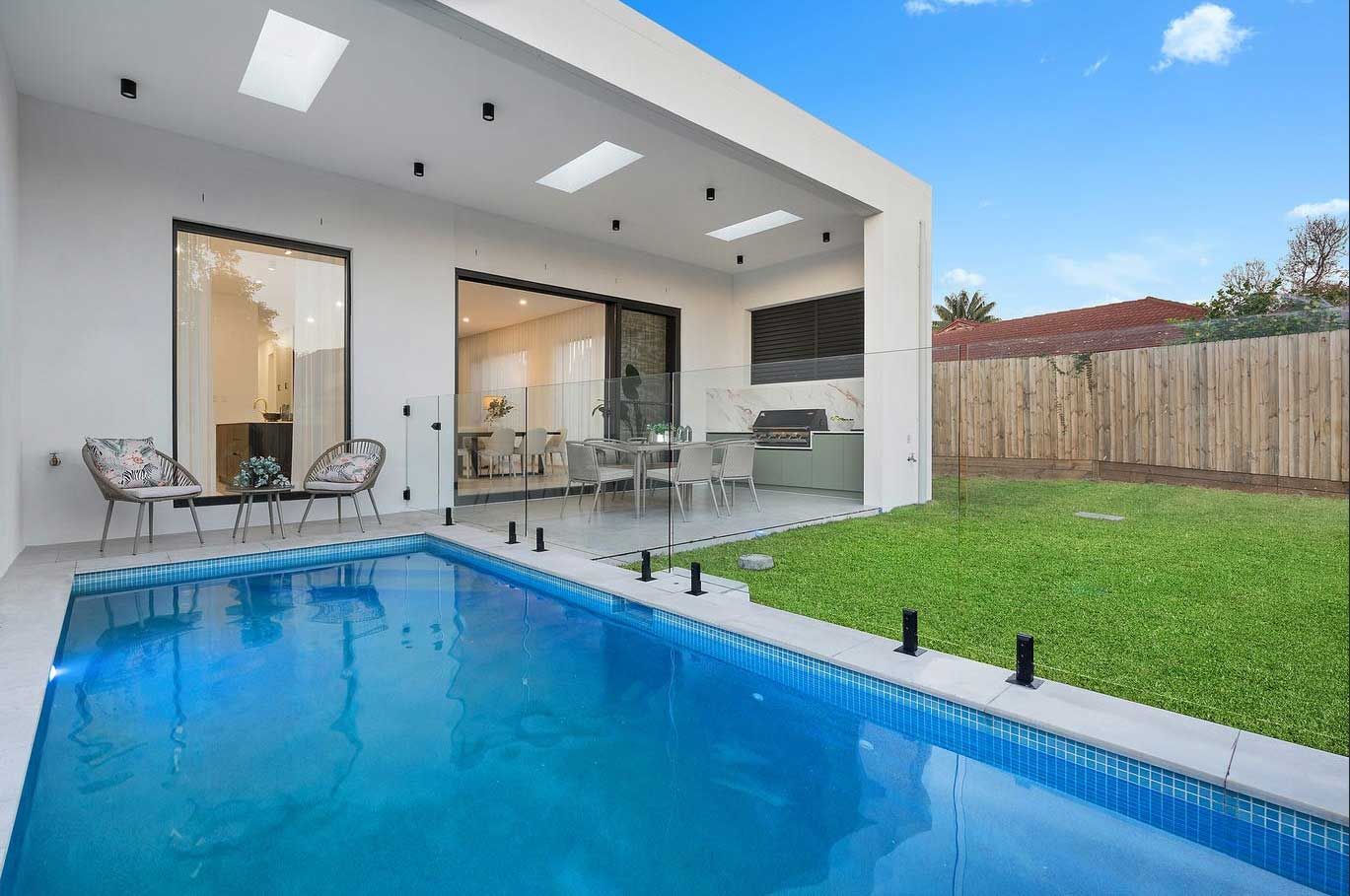 Backyard with pool, outdoor dining area, and grass lawn. Bright blue pool, white walls, and clear sky.