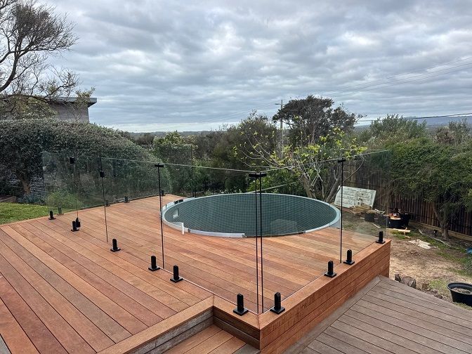 Deck with a round tub surrounded by a glass and black railing, overlooking a scenic landscape under a cloudy sky.