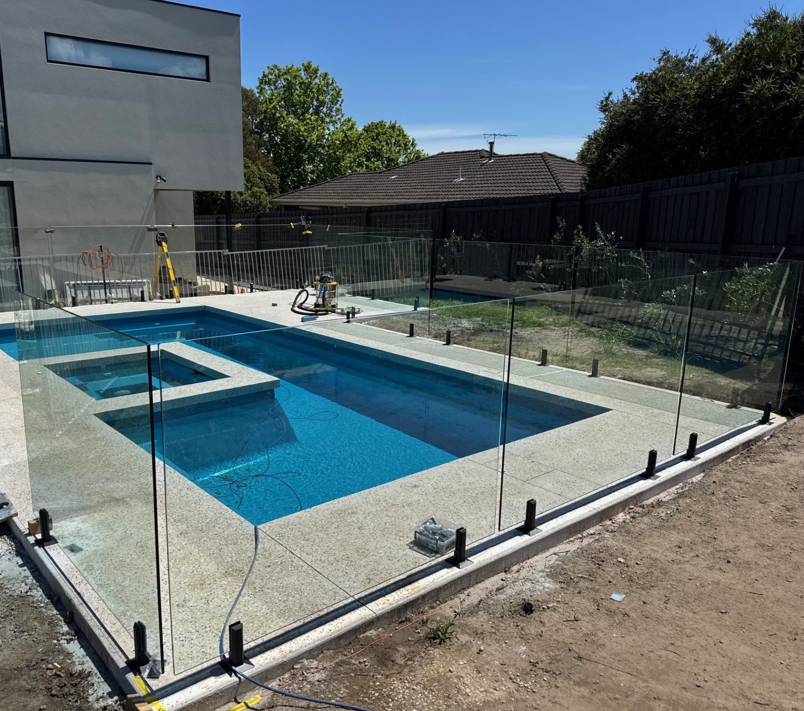 Swimming pool with glass fence, concrete deck, and house in the background on a sunny day.