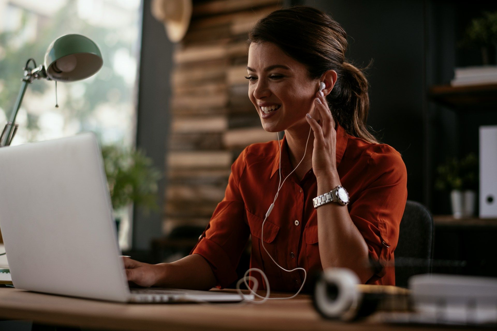 Woman in orange shirt at laptop, smiling, with earbuds. Desk, lamp, and window visible.