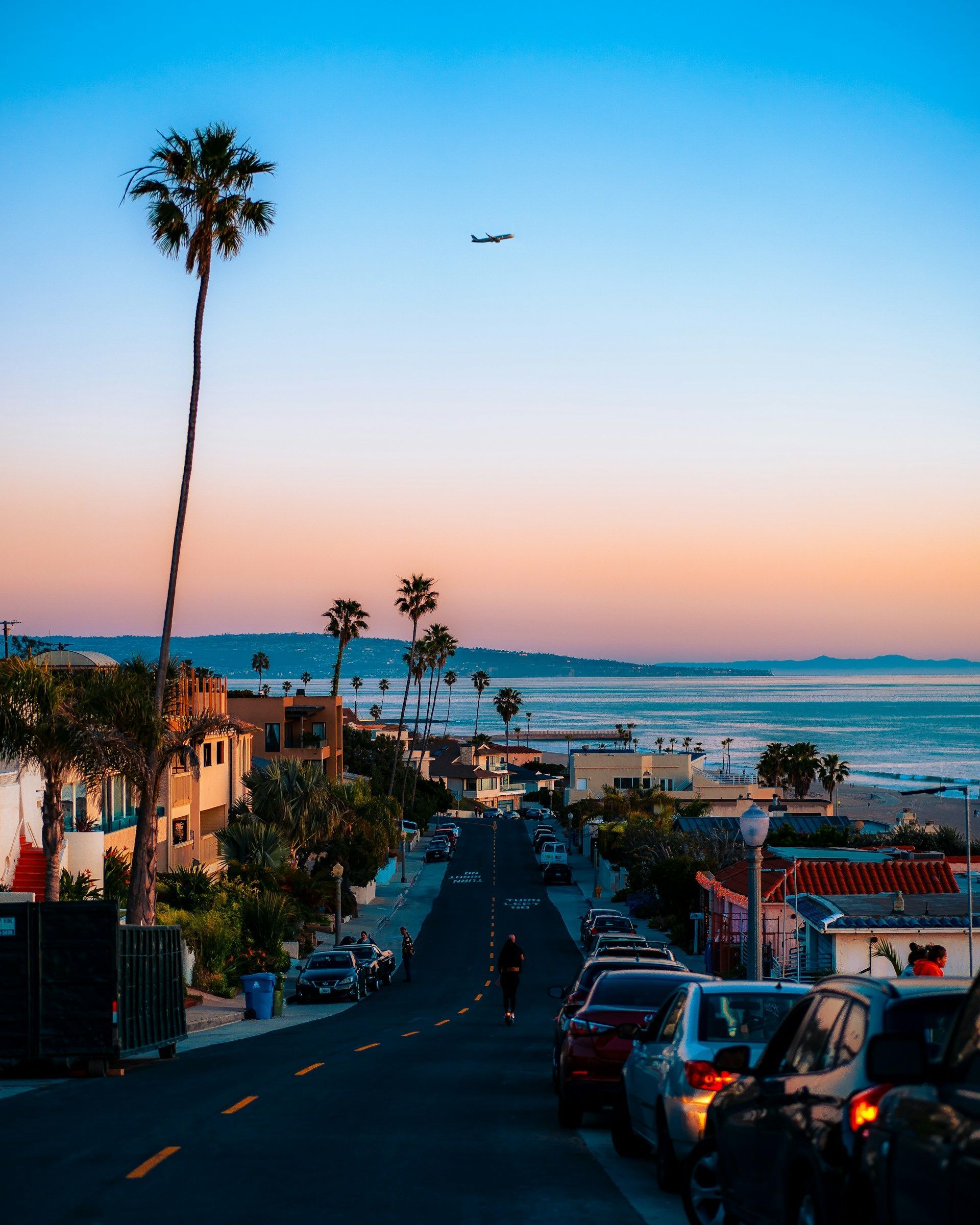 Street view towards the ocean at sunset with palm trees, cars, and a plane.