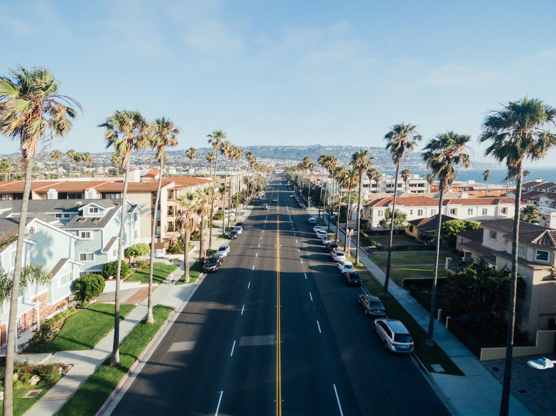 Street lined with palm trees and buildings, leading to the ocean under a clear blue sky.