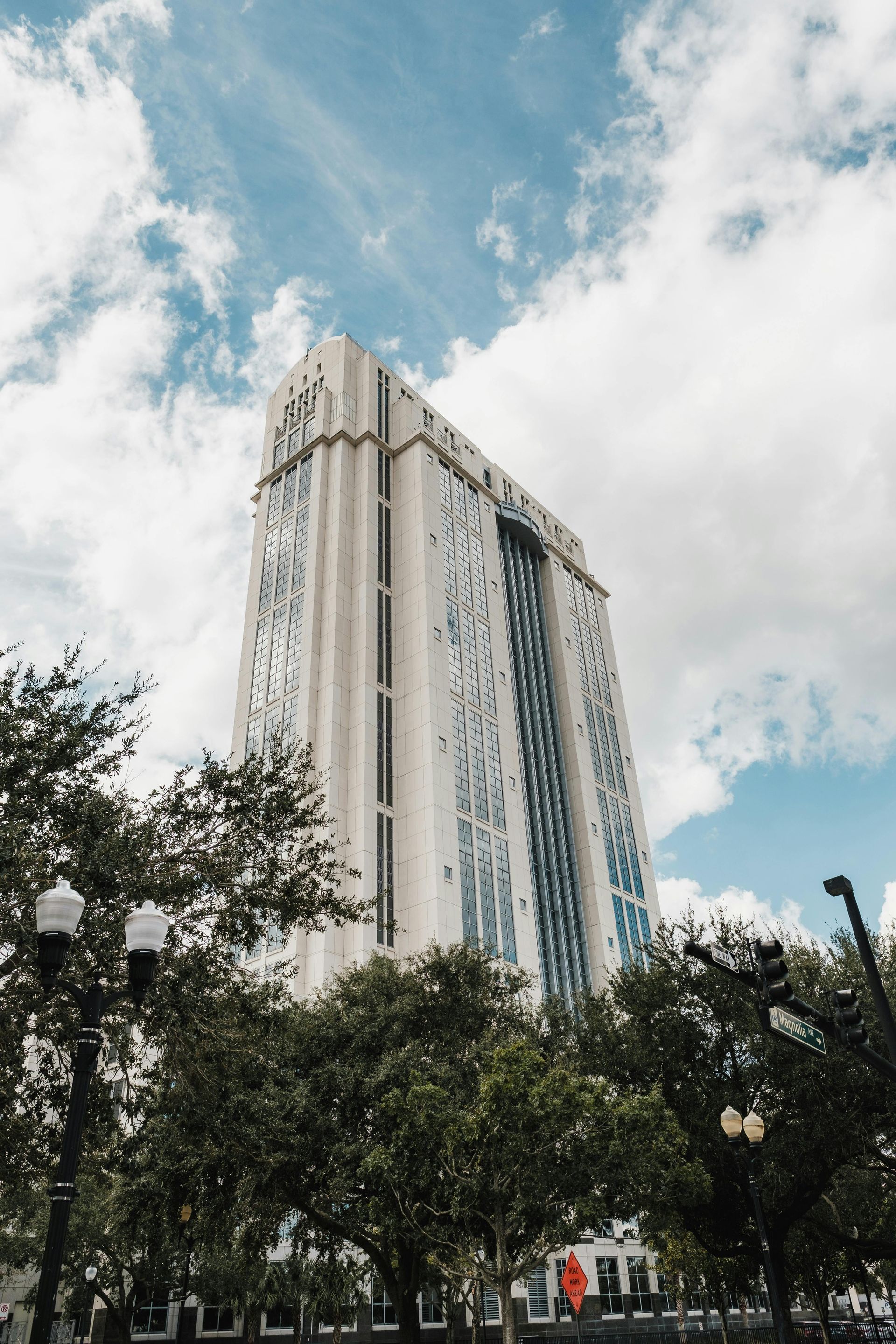 Tall, white skyscraper against a cloudy blue sky, viewed from street level. Lush green trees frame the building.