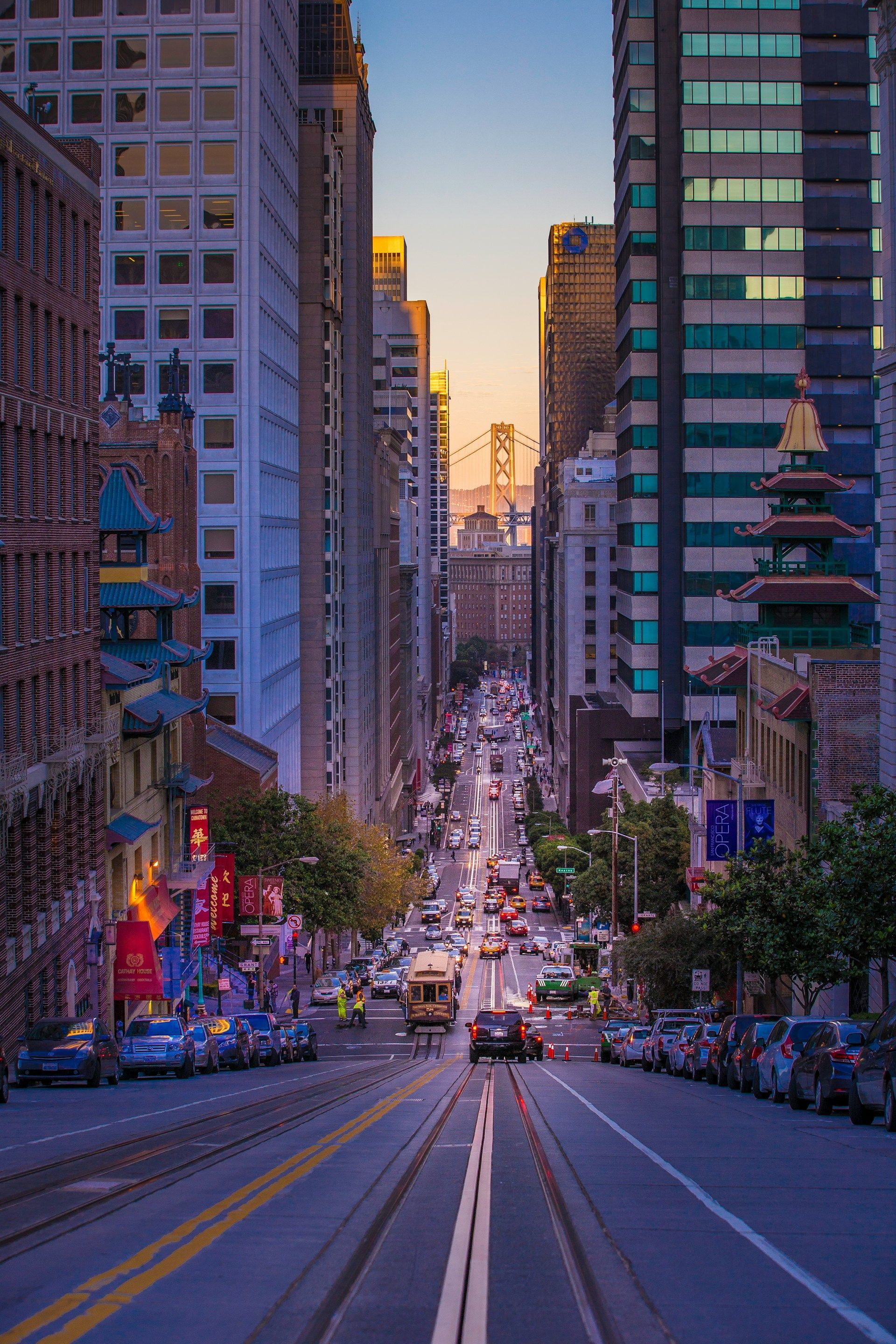 Steep San Francisco street scene with cars, cable car, and city buildings; Bay Bridge visible in distance.