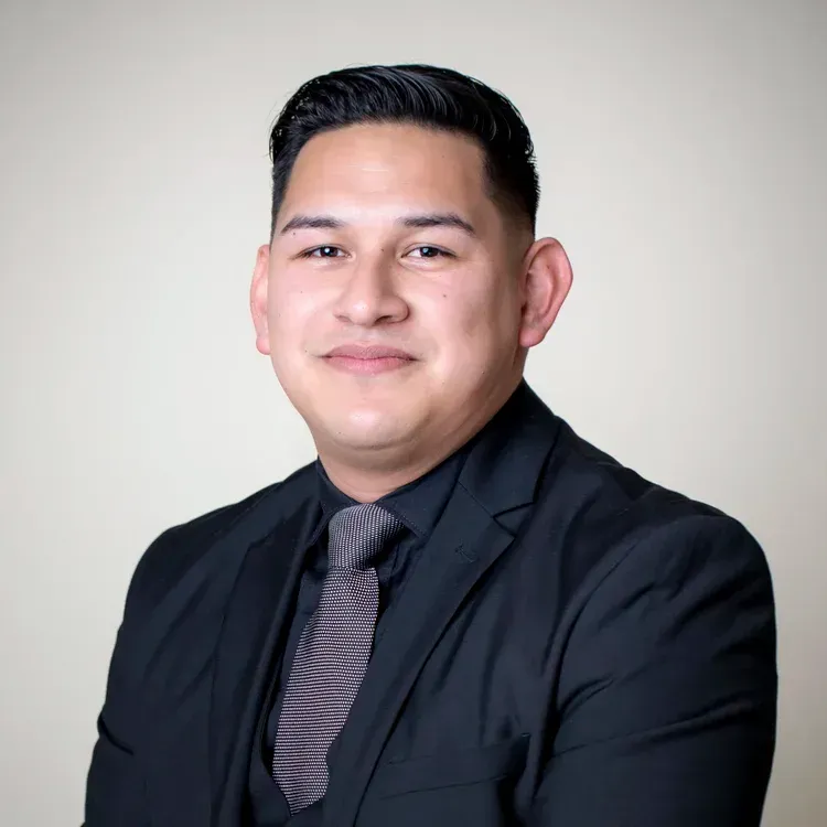 Man in a black suit with a striped tie, smiling in front of a neutral background.