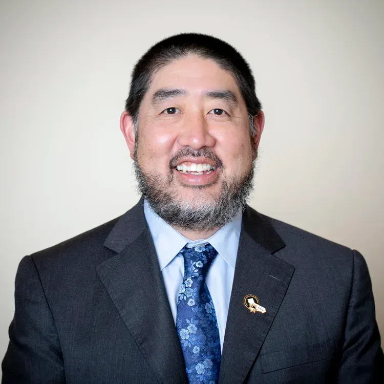 Man in a suit smiles, wearing a patterned blue tie and a lapel pin, against a neutral background.