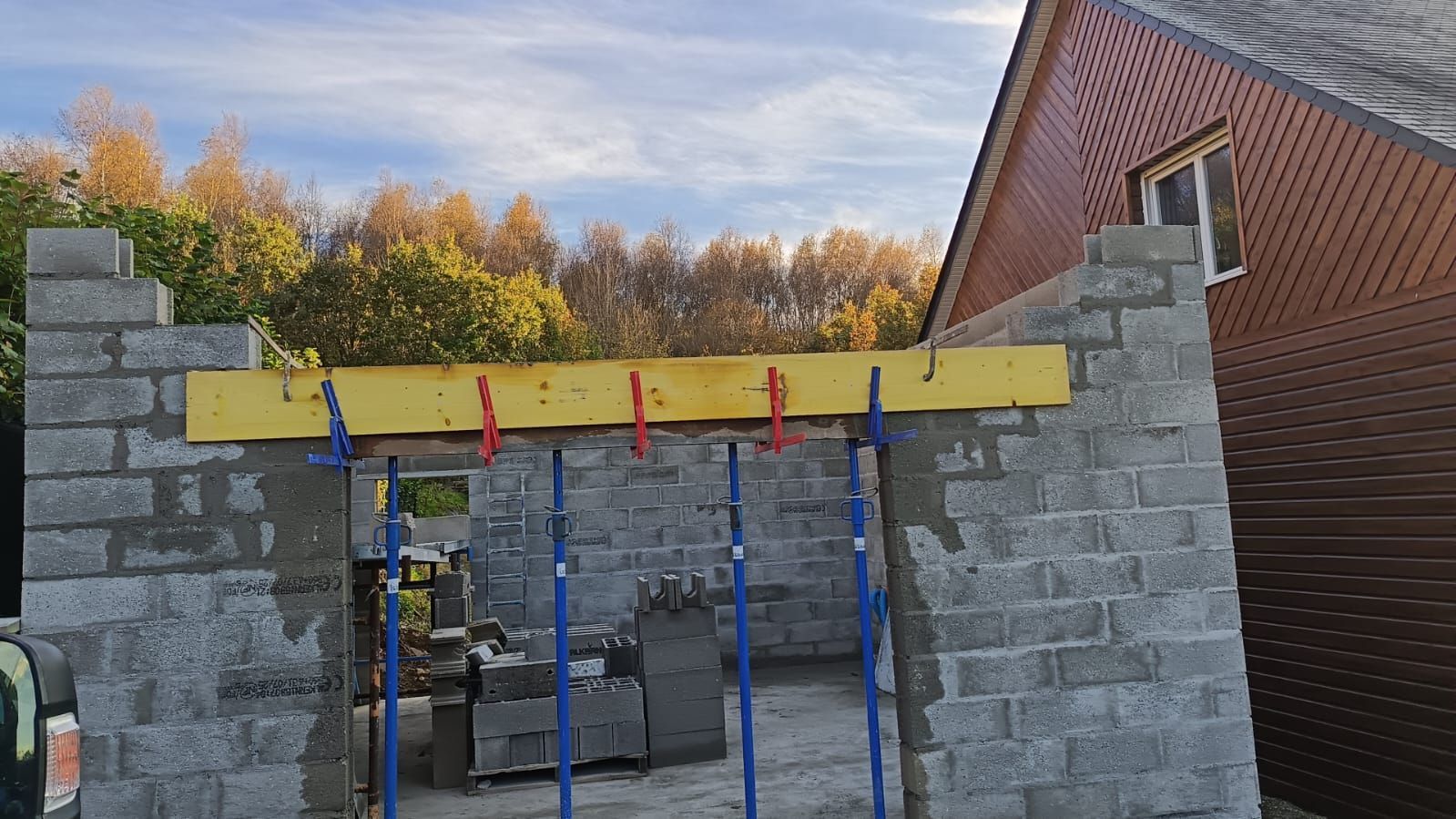 Mur en briques partiellement construit avec poutre jaune et supports bleus/rouges sur un chantier extérieur