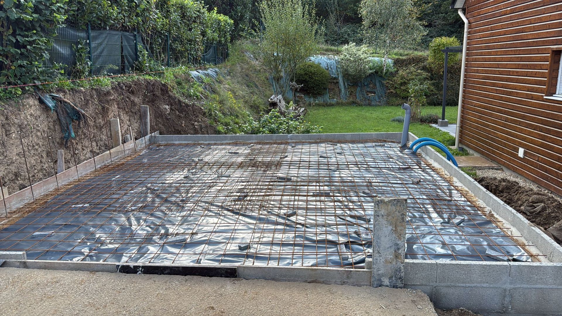 Dalle de béton nue au fond d'un jardin, à côté d'une maison en briques et d'un mur de soutènement.