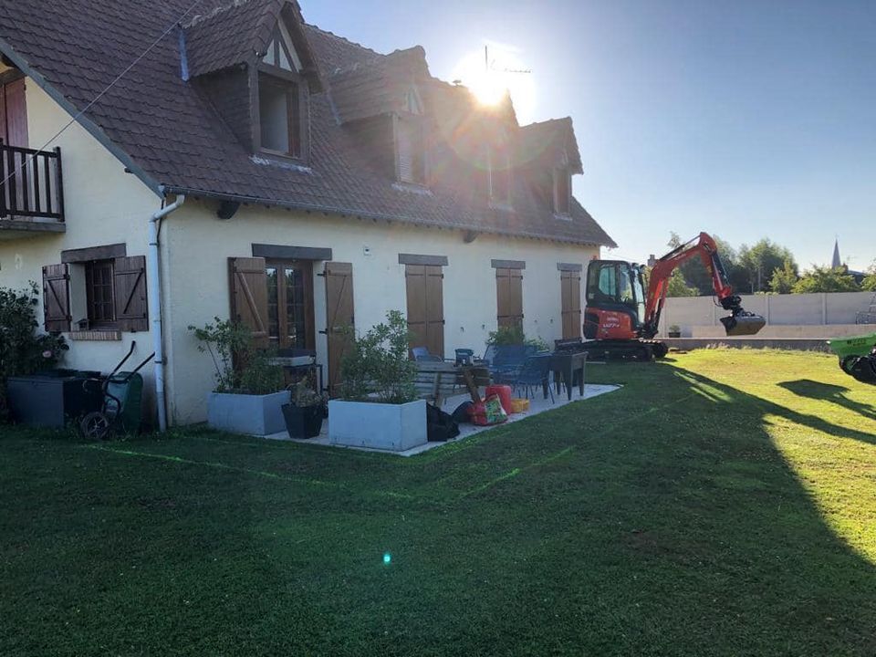 Maison avec parterres de fleurs et pelouse, pelleteuse en activité près d'un mur blanc sous un soleil éclatant