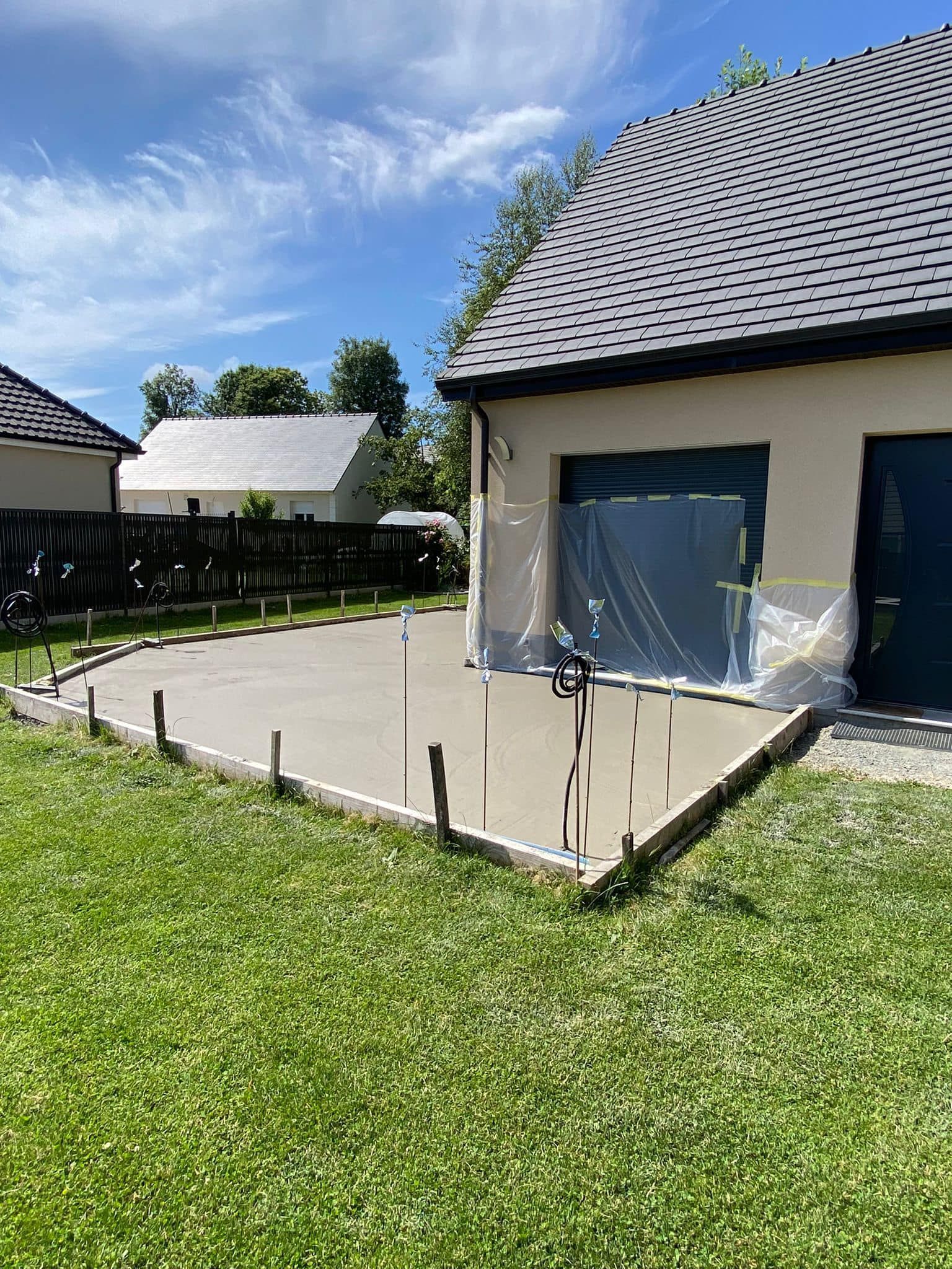 Terrasse arrière attenante à une maison, avec dalle de béton, pelouse et ciel bleu.