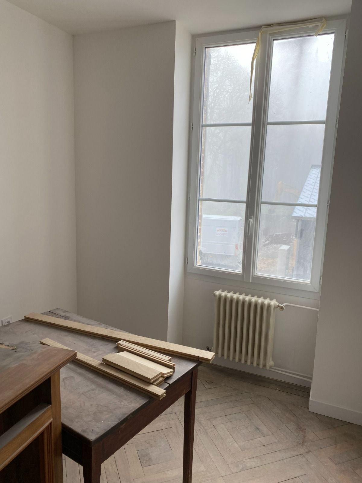 Pièce spartiate avec une fenêtre et un radiateur, ainsi qu'une table sur laquelle reposent des planches de bois.