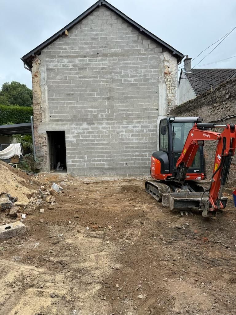 Chantier de construction avec un bâtiment en béton inachevé et une excavatrice orange sur un sol en terre battue