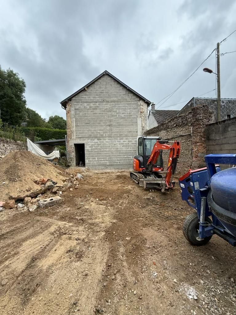 Chantier de construction avec allée de gravier, bâtiment en pierre, excavatrice et scooter bleu sous un ciel nuageux