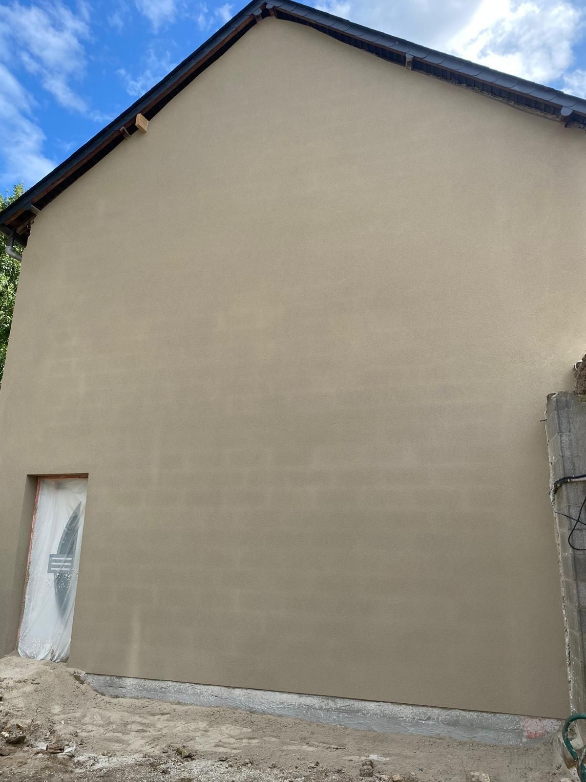 Mur latéral d'une maison beige uni avec une porte blanche et un toit en pente sous un ciel bleu