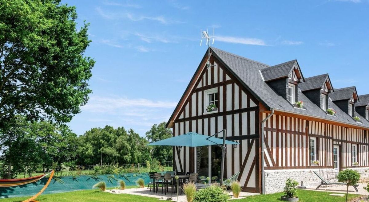Maison moderne avec poutres apparentes au bord d'une piscine turquoise, parasol, pelouse et arbres sous un ciel bleu.