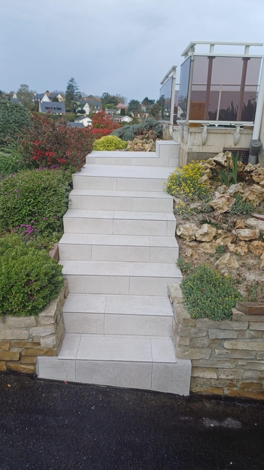 Des marches en pierre mènent à un chemin de jardin bordé d'arbustes, de fleurs et d'une terrasse avec balustrade en verre.