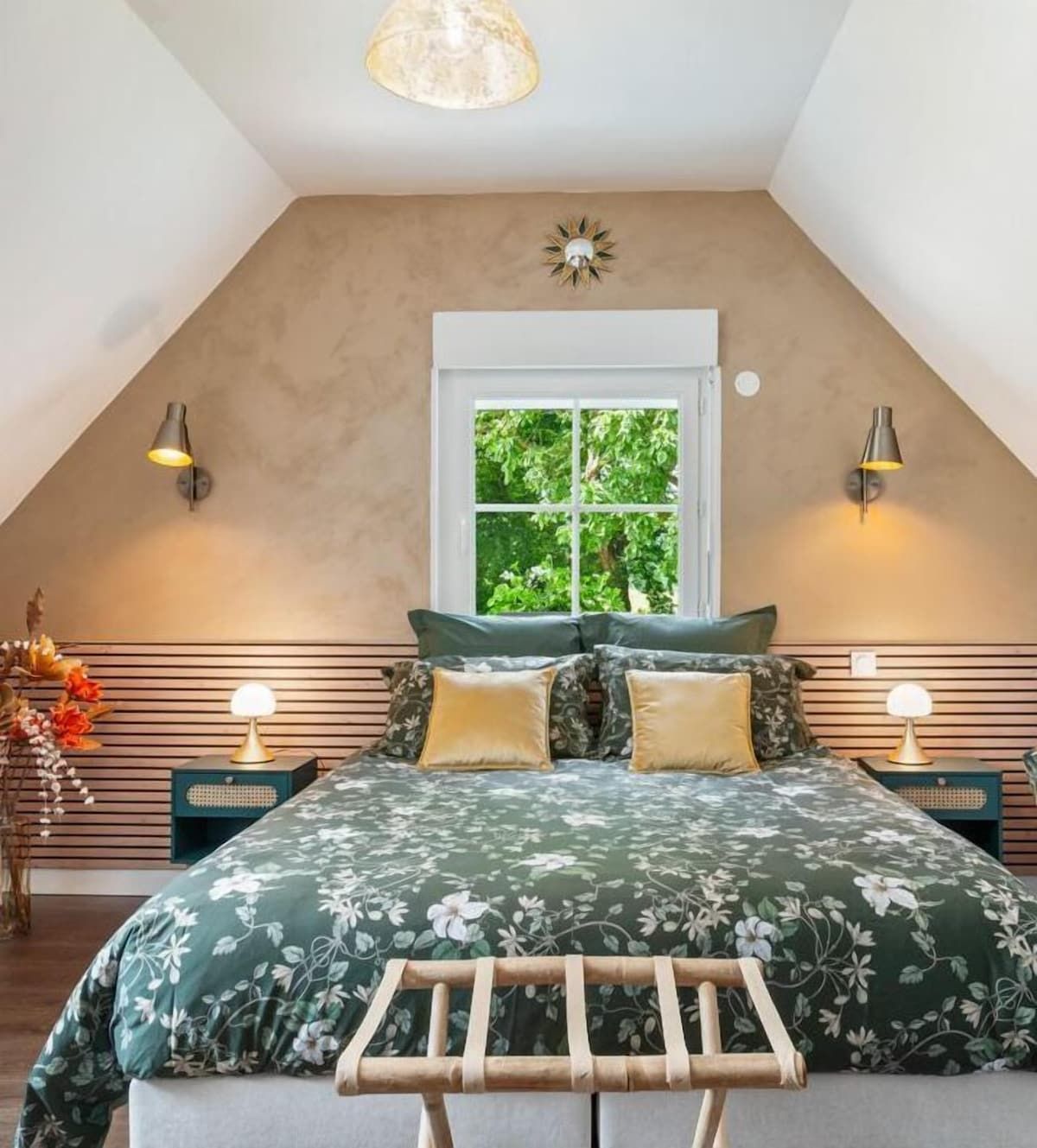 Chambre confortable avec un couvre-lit vert à motifs floraux, une tête de lit en bois, une fenêtre et des appliques murales.