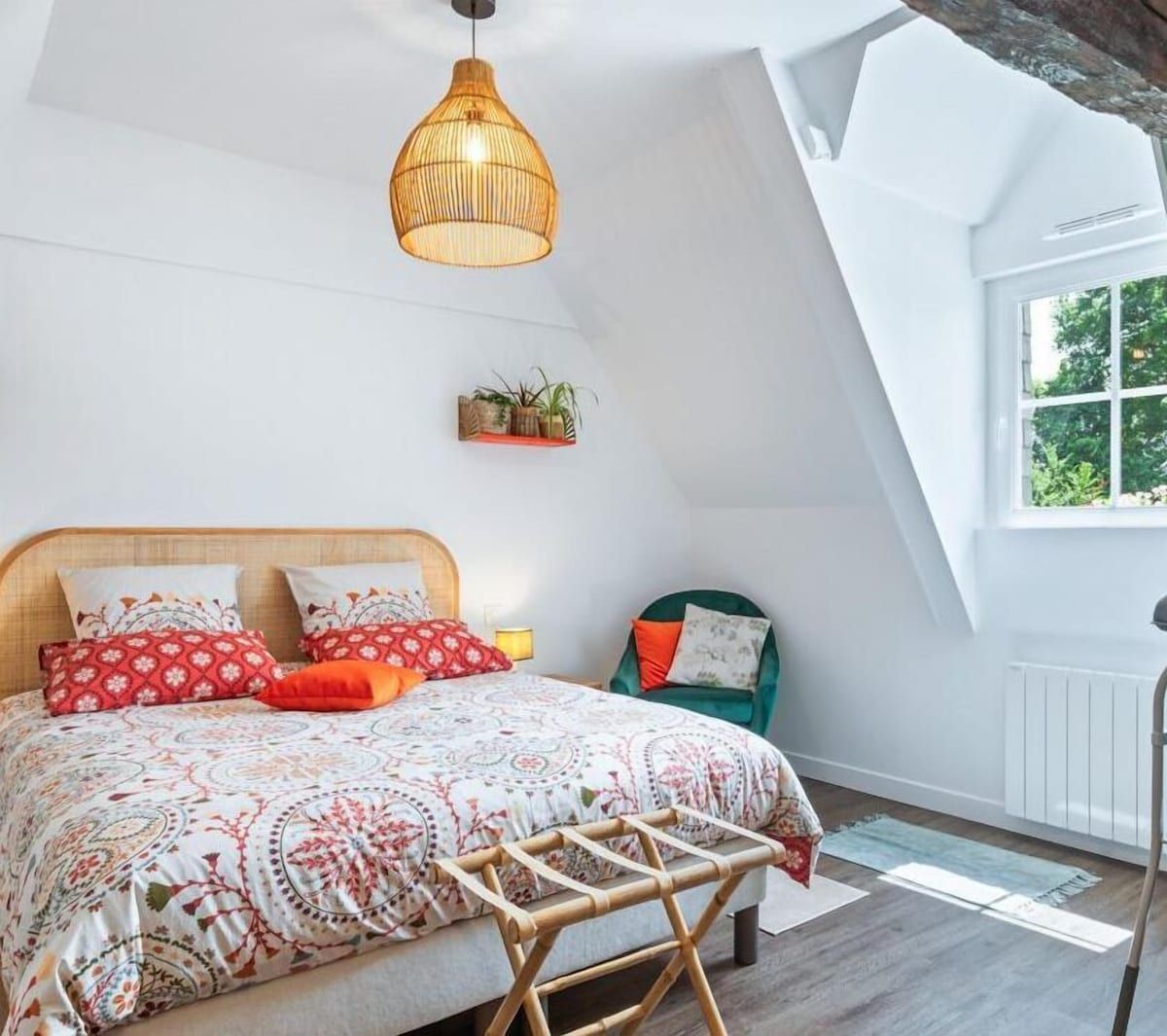 Chambre lumineuse avec un lit à motifs, une suspension en osier, une petite chaise et une fenêtre ensoleillée sous un plafond mansardé