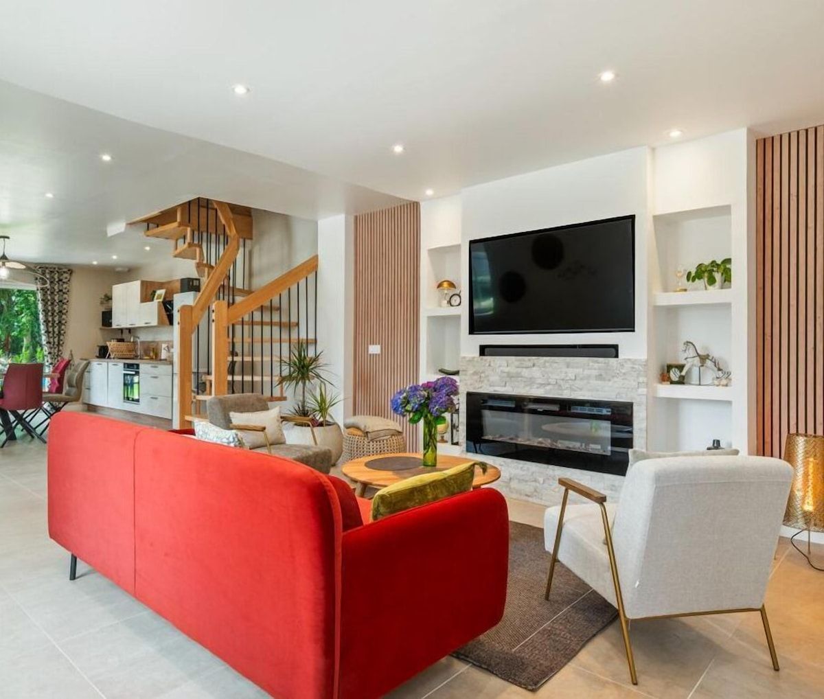 Salon moderne et lumineux avec canapé rouge, fauteuil blanc, télévision murale et escalier ouvert.