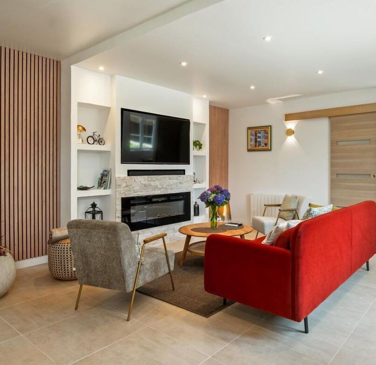 Salon moderne avec canapé rouge, fauteuil gris, télévision murale, cheminée et table basse