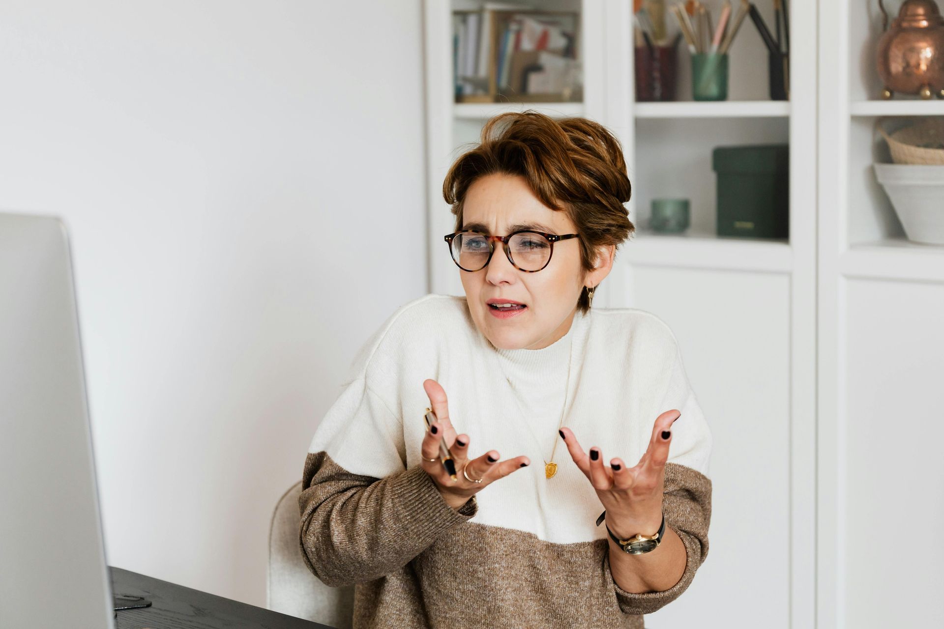 Woman with glasses, hands raised in an exasperated gesture, looking at a laptop, neutral background.